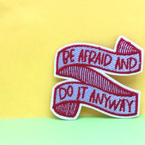 Könnte beinhalten: Lila und weiß gestickter Patch mit dem Text "Hab Angst und tu es trotzdem" in rotem Garn.
