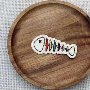 Può includere: Una toppa in tessuto a forma di scheletro di pesce, con base color crema e contorno nero. Le costole colorate sono rosse, gialle, turchesi e verdi. La toppa è posizionata su un vassoio rotondo in legno con finitura marrone scuro.