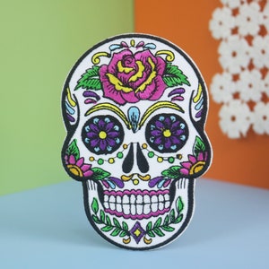 Könnte beinhalten: Ein weißer gestickter Zucker Schädel Patch mit bunten Blumen, grünen Blättern und einer rosa Rose. Der Schädel hat lila und gelbe Details um die Augen und den Mund.