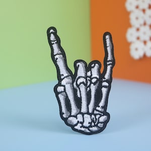 Puede incluir: Un parche bordado en blanco y negro de una mano de esqueleto haciendo el gesto de rock and roll.