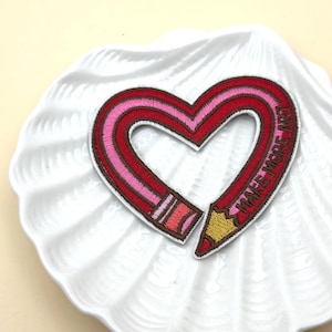 Pode incluir: Remendo bordado em forma de coração, desenhado como um lápis. O lápis é vermelho e rosa com uma ponta amarela. As palavras "MAKE MORE ART" são bordadas na lateral. O remendo está em um prato branco em forma de concha.