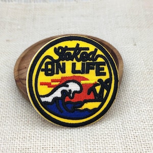 May include: A circular embroidered patch featuring a yellow background with a black border. The design depicts a red sunset, a white ocean wave, and black palm tree silhouettes. The text "Stoked ON LIFE" is embroidered in black across the top.
