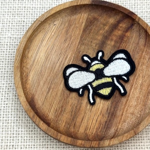 Op de afbeelding: Geborduurde bijenpatch met zwarte omtrek, witte vleugels en geel en zwart lichaam, op een ronde houten schaal. De schaal heeft een natuurlijke houtnerf en staat tegen een neutrale stoffen achtergrond.