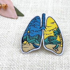 Puede incluir: Un parche bordado con forma de pulmones humanos. El pulmón izquierdo presenta un paisaje montañoso azul con un sol y una tienda de campaña sobre suelo arenoso. El pulmón derecho presenta un cielo amarillo con una palmera, una gran ola y una silla de playa.