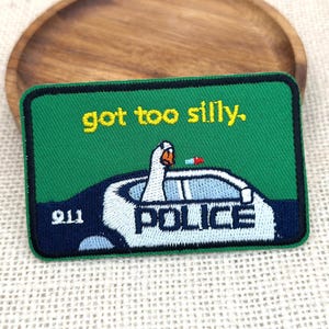 Peut inclure: Un écusson vert avec le texte "got too silly." présente une voiture de police avec une oie au volant. Le mot "POLICE" est affiché sur la voiture et le numéro "911" est dans le coin.