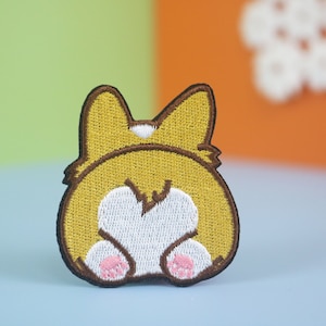 Könnte beinhalten: Ein gestickter Aufnäher, der das Hinterteil eines Corgis zeigt, das dem Betrachter den Rücken zukehrt. Der Aufnäher ist braun, gelb und weiß, mit einem braunen Rand und aus weichem, plüschem Stoff.