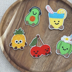 Puede incluir: Un conjunto de parches bordados con frutas y bebidas sonrientes. Incluye un aguacate con auriculares, una piña, cerezas, un cóctel y una bebida de coco. Sobre una bandeja de madera.