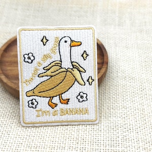 May include: Embroidered patch featuring a goose in a banana peel costume. The rectangular patch has a white background with gold trim, the goose is yellow and white with orange feet and beak. Text reads "I'm not a silly goose, I'm a banana."