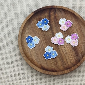 Może przedstawiać: Haftowane naszywki z kwiatami w odcieniach niebieskiego, różowego i białego, ułożone na drewnianym talerzu. Każda naszywka przedstawia skupisko małych kwiatów z żółtymi środkami i białymi obwódkami. Naszywki umieszczone są na neutralnym tle z juty.