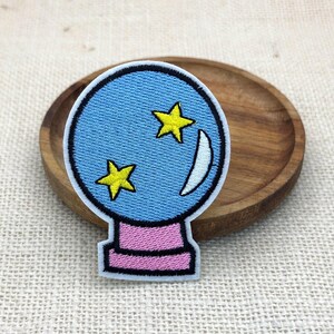 May include: An embroidered patch featuring a circular blue crystal ball design with two yellow stars and a white crescent moon. The crystal ball sits on a pink rectangular base, and the entire shape is outlined in a thick black border.