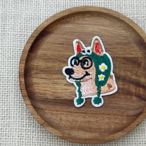 May include: Embroidered patch featuring a cartoon dog wearing a green hat with white daisy accents and glasses. The dog has a tan and white face with a black nose and smiling mouth. The patch is on a round wooden tray.