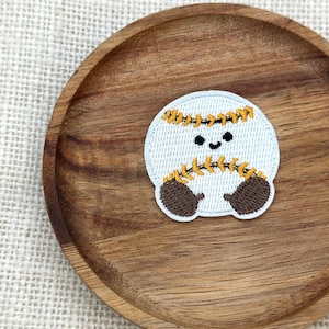 Puede incluir: Un parche con temática de béisbol con una cara sonriente, con un fondo azul claro, guantes marrones y costuras amarillas. El parche está colocado sobre un plato de madera con un fondo de tela clara.