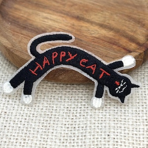 May include: Black embroidered patch in the shape of a cat with the words "HAPPY CAT" in red. The cat has white paws and a white tip on its tail. The cat's eyes are white with red accents.