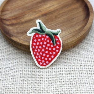 May include: Embroidered strawberry patch with a red body and green leaves. The patch has a white border and is placed on a wooden plate. The strawberry is covered in small pink dots.