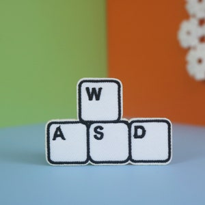 Puede incluir: Parches de tela blanca con forma de teclas de ordenador con las letras "W", "A", "S" y "D" en bordado negro. Los parches están dispuestos en forma de pirámide.
