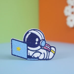 Può includere: Una toppa ricamata con un astronauta dei cartoni animati che indossa una tuta spaziale bianca e un casco blu, seduto davanti a un laptop blu con una stella gialla sullo schermo.