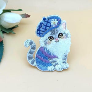 May include: Embroidered patch of a fluffy gray and white cat wearing a blue beret with a white flower. The cat has blue and purple stripes and is sitting with its tail curled. The patch is on a light beige background.