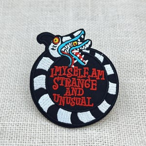 May include: A black and white embroidered patch with the text "I MYSELF AM STRANGE AND UNUSUAL" in red. The patch features a cartoonish creature with a blue and white head and a red tongue, surrounded by a black and white striped border.