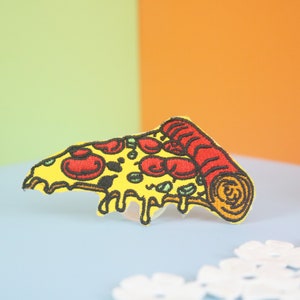 May include: Embroidered pizza slice patch with yellow crust, red pepperoni, and green olives. The melted cheese drips down the side. The patch is on a blue surface with a colorful background.