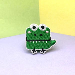 May include: A green embroidered alligator patch with a white border. The alligator has white eyes, a black mouth, and small pink cheeks. The patch is on a white surface with a green and yellow background.