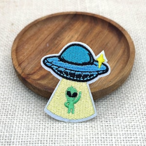 May include: Embroidered patch featuring a blue UFO with a green alien waving from the bottom. The UFO has a black outline and a yellow star. The patch is on a light yellow background with a white border.