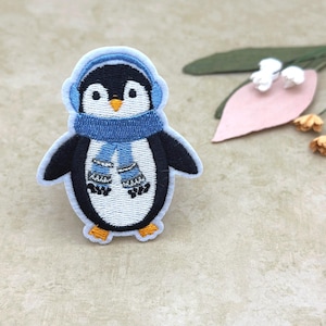 May include: Embroidered patch of a cartoon penguin wearing blue earmuffs and a scarf. The penguin has a white belly, black wings, and orange feet and beak. The patch is outlined in white.