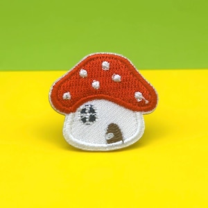 Può includere: Toppa ricamata a forma di fungo con cappello rosso a pois bianchi. Il fungo ha una base bianca con una piccola finestra e una porta marrone. Su sfondo giallo e verde.