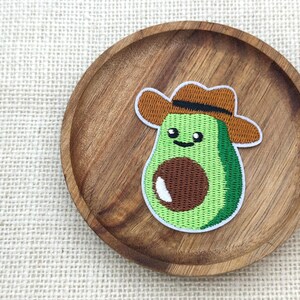 以下が含まれることがあります： 茶色のカウボーイハットをかぶった、笑顔のアボカドの刺繍パッチ。アボカドは緑色で、茶色の種と白いハイライトがあります。パッチは丸い木製のプレートの上にあります。