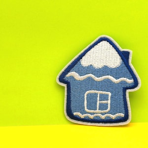 May include: Embroidered patch in the shape of a blue house with a white roof and window. The house has a white outline and is set against a bright yellow and green background.