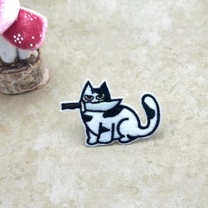 May include: Embroidered patch featuring a black and white cat holding a knife. The cat has a stern expression. The patch is white with black details and a black outline. A mushroom decoration is in the background.