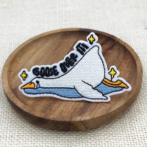 May include: An embroidered patch featuring a white goose lying down on a blue surface with yellow star accents. The text "GOOSE OVER IT" is stitched in black letters above the goose. The patch has a white border and is shaped like the goose.