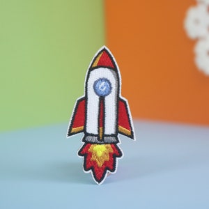 May include: An embroidered rocket patch in red and white with a blue window and yellow flames. The patch is on a light blue background.