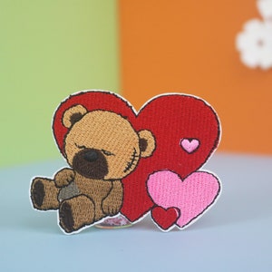 May include: Embroidered iron-on patch featuring a brown teddy bear with stitched eyes and a red heart behind it. There are two smaller pink hearts below the bear and a smaller red heart on the large red heart.