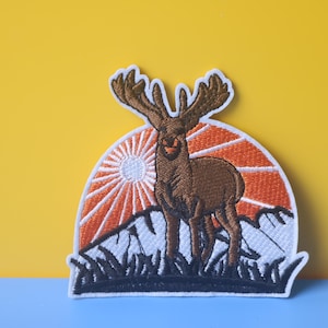 以下が含まれることがあります： 大きな角を持つ茶色のヘラジカが、オレンジと白の夕焼けを背景に刺繍されたパッチ。 ヘラジカは、白と黒のディテールを持つ山脈の前に立っています。