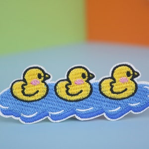 Può includere: Tre paperelle di gomma gialle galleggiano in una piscina blu. Le paperelle hanno le guance rosa e gli occhi neri. La piscina è circondata da cuciture bianche.