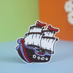 Può includere: Una toppa ricamata raffigurante un veliero con scafo marrone scuro, finiture viola e vele bianche. La nave presenta tre alberi con bandiere rosse sulla sommità e piccoli dettagli circolari bianchi lungo il fianco dello scafo, bordata di bianco.