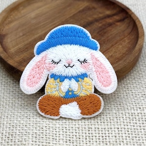 May include: An embroidered patch featuring a white rabbit with closed eyes, pink inner ears, and rosy cheeks. The rabbit wears a blue hat, a blue and yellow patterned shirt, and sits in a cross-legged position with brown legs and a white tail.