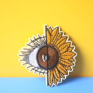 Puede incluir: Un parche de tela blanca bordado con un girasol amarillo con un centro negro. El girasol está medio cubierto por un ojo blanco con un iris marrón y pestañas negras.