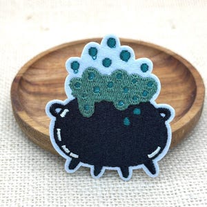 Könnte beinhalten: Ein gestickter Aufnäher, der einen schwarzen Kessel mit hellblauem Umriss zeigt. Der Kessel enthält sprudelnde grüne Flüssigkeit mit dunkelgrünen kreisförmigen Akzenten und hellblauen Blasen, die über den Rand steigen, alles auf weißem Rand.