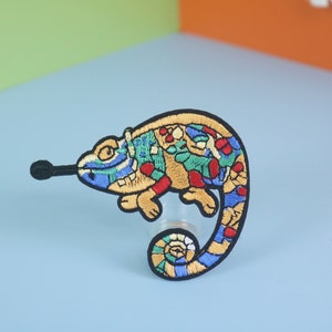 May include: A colorful embroidered iron-on patch featuring a chameleon with a blue, green, red, and yellow design.