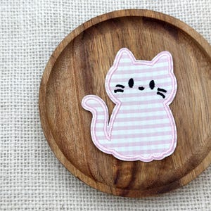 以下が含まれることがあります： 黒い顔の特徴を持つピンクと白のギンガムチェックの猫のパッチが、木製のコースターの上にあります。パッチは白い縁取りのある刺繍アップリケです。コースターは木製で、自然な仕上がりです。
