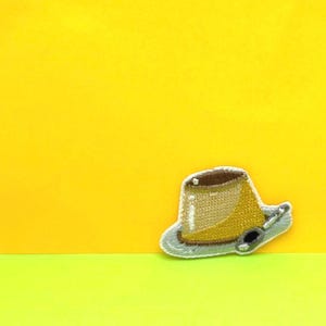 May include: A brown and yellow embroidered iron-on patch of a pudding cup with a spoon.