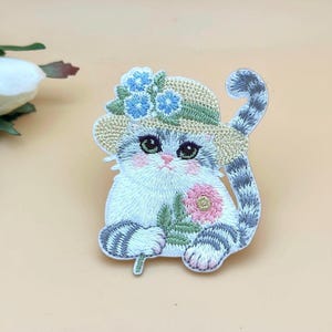 May include: Embroidered patch featuring a charming cat wearing a straw hat decorated with blue flowers. The cat holds a pink and green flower, with a striped tail and paws. The patch is white, grey, and pink, with green eyes.