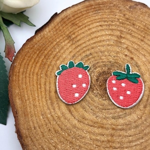 May include: Two embroidered strawberry patches. Each patch features a red strawberry with white seeds and a green stem. The patches are outlined in white and are set against a wooden background.