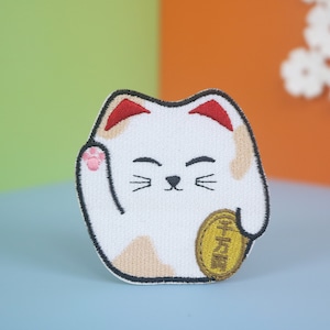 May include: Embroidered patch featuring a white Maneki-neko cat with red ears and a raised paw. The cat holds a gold coin with Japanese characters. The patch has a black border and is set against a colorful background.
