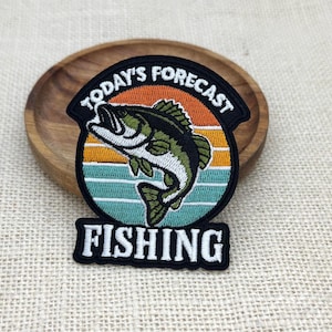 May include: A circular embroidered patch featuring a green and white fish jumping in front of a sunset background with orange, yellow, and teal stripes. The text reads TODAY'S FORECAST at the top and FISHING at the bottom in white letters.
