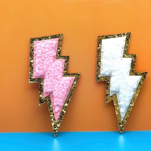 May include: Two lightning bolt shaped patches with gold glitter trim. One patch is pink and the other is white.