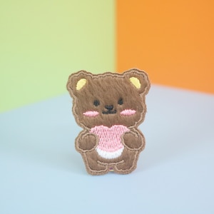 Könnte beinhalten: Gestickter Aufnäher eines braunen Teddybären mit einem rosa Herz. Der Bär hat gelbe Ohren und rosa Wangen. Der Aufnäher ist zum Aufbügeln oder Aufnähen auf Stoff gedacht. Der Hintergrund ist ein Farbverlauf aus Grün, Blau und Orange.