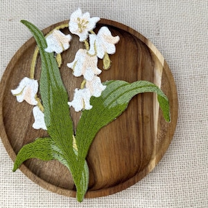 May include: An embroidered patch featuring a lily of the valley plant with white bell-shaped flowers and green leaves. The patch is placed on a round, dark brown wooden tray. The embroidery uses white, light peach, and various shades of green thread.
