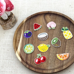 以下が含まれることがあります： 木製のトレイには、スイカのスライス、桃、パイナップル、レモンなど、カラフルな刺繍フルーツパッチが並んでいます。背景には赤と白のキノコの装飾があります。パッチは、衣類やアクセサリーの装飾に最適です。